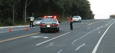 A photograph depicting two police officers directing traffic on a roadway. The officers are wearing reflective vests, and their patrol car is parked with its roof lights active in the middle of the road at a turn junction. Several orange traffic cones are blocking the left lanes of the road, and the officers appear to be directing traffic onto an alternate route.