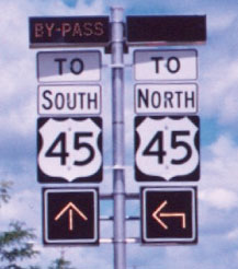 A photograph of a signpost with two columns of signs, one column mounted on either side of the signpost. Both columns are topped by rectangular, non-illuminated signs that would read “Bypass” if lit. Beneath that, the lefthand column reads “To south 45” on a white sign with black lettering, while a similar sign on the right reads “To north 45.” At the bottom of each column is a square changeable arrow sign, appearing with white lettering on a black background. The lefthand sign points straight up, while the righthand sign makes a 90 degree turn to point left.