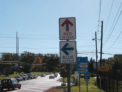 A photograph of a signpost perhaps 50 feet before a four-way intersection with a traffic light. The sign is mounted on the right side of the road. The signpost bears two rectangular sign, one above the other. The top sign is white with red lettering, and has an arrow pointing straight up with the words “Red detour” beneath the arrow. The lower sign is white with blue lettering and has an arrow pointing to the left above the words “Blue detour.”
