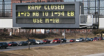 Changeable message sign displaying message to divert traffic to an alternate route. The sign is hanging over a congested freeway.