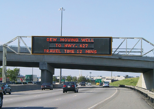 Variable message sign above highway that states "QEW moving well to hwy 427 travel time 12 mins."