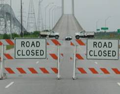Photo of a road leading to a bridge. The road has been blocked by Road Closed warning signs, two trucks, and a police cruiser.