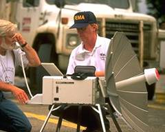 Photo of a man using a satellite phone that is hooked up to a portable satellite dish.  A FEMA employee looks on.
