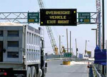 photo of a truck passing under an overhead roadway sign that reads "overheight vehicle exit right"