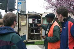photograph of four engineers standing in front of an open controller cabinet. One person is pointing to the hardware in the cabinet while discussing how the hardware works. The simulation allows for fine-tuning of the signal priority strategies prior to implementation in the field.