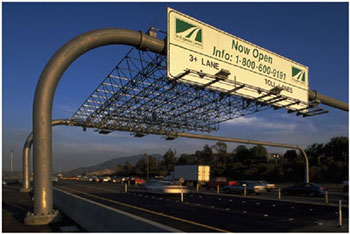 Photo. An overhead antenna apparatus on a busy highway. Overhead antennas like this one are used to read transponders on the vehicles below, allowing tolls to be paid without the cars having to stop.