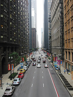 Photo. View of cars driving one way down a four-lane city street with tall buildings on either side. The fourth lane furthest to the right is an HOV lane.