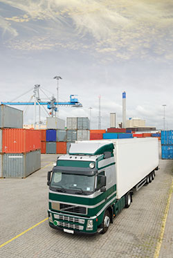 Photo. A large truck in a freight yard at a port facility, surrounded by large shipping containers, with a crane in the distance.
