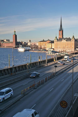 Photo. A view of the city of Stockholm. In the background is the city skyline on the water; in the foreground traffic is flowing freely over a bridge.