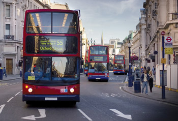 Photo. A street-level view of four double-decker buses travelling up a central London street with no other traffic.