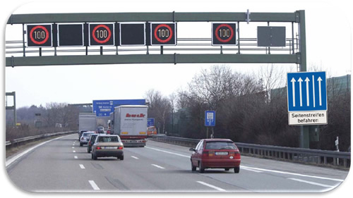 Figure 28. Photo. Right Shoulder Use with Speed Harmonization—Germany. Photo showing temporary shoulder use operational with speed harmonization on an urban freeway in Germany. An overhead sign gantry indicates reduced speed limits for each lane, including the shoulder, and sign on the shoulder indicates the shoulder is open to traffic.