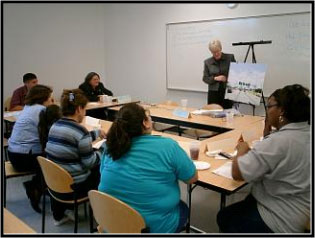 Photo of individuals sitting around U-shaped table watching a presentation by an individual standing at the front of the room with an easel display.
