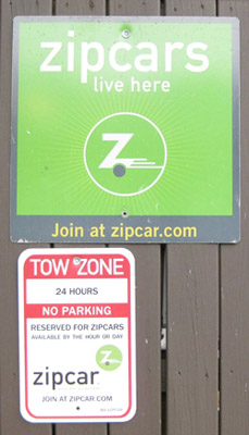 Photo of a green parking sign stating "Zipcars live here" above a Tow Zone sign stating "Reserved for Zipcars."