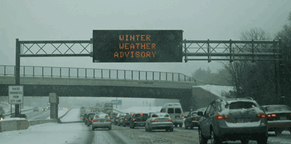 Picture showing traffic in the snow along a limited access highway. There is a dynamic message sign over the roadway that says “winter weather advisory.”