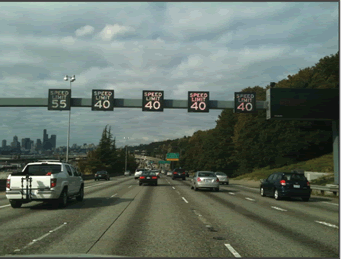 A photograph of interstate lanes with an overhead truss indicating a speed limit over each travel lane, the four right lanes are signed at 40 mph and the left lane is signed at 55 mph.