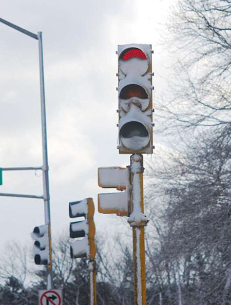 This is a photograph of a traffic light facing the camera, with the red light lit. There are two traffic lights behind it, facing to the left. Trees, a tall overhead light pole and a No Right Turn sign are also in the background. There is snow accumulated on the surfaces of the traffic light, including the interior surface of the lights. As a result the red light is barely visible.