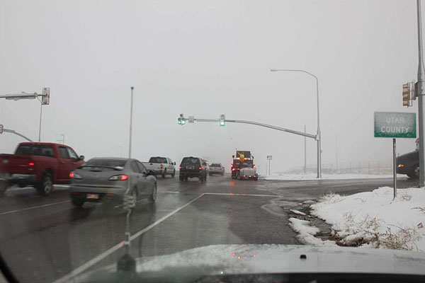 Figure 3. Cleaning Lenses with Telescoping Pole (M. Taylor). This is a photograph of a street scene in snowy weather, near a traffic intersection. There is snow on the ground to the right side of the street, a snow-dusted sign reading Utah County on the right, and the roads appear wet. Cars are traveling away from the camera through the intersection, with the traffic lights showing green. On the right shoulder on the far side of the intersection, there is a road-work vehicle with warning lights stopped alongside the overhead traffic lights. A faint telescopting pole can be seen extending from the truck up to the rightmost traffic light.