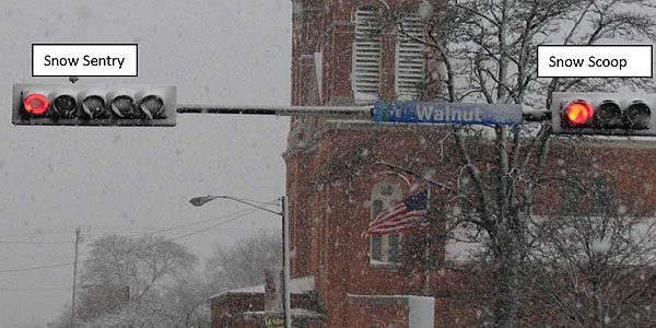 Figure 6. Snow Sentry vs. Snow Scoop (D. Hansen). This is a photograph of a snowy intersection with a brick building in the background and the overhead traffic poles with horizontal lights in the foreground. The traffic light to the left is marked with an arrow with the words Snow Sentry. The red light is lit, and has a strong red outline and a dimmer center. The right traffic light is marked with an arrow with the words Snow Scoop. The red light on this side is also lit, but glows with more brightness.
