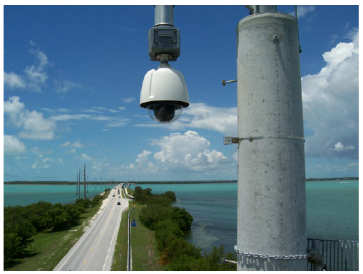 A CCTV camera mounted on a post with a view of a long bridge over a body of water.