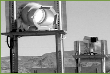 A detailed photo of a monitoring station used to capture the license plate information of cars traveling through the work zone. The monitoring station consists of two small pole-mounted signs with a light mounted on the back of one sign, and a camera mounted on the back of the other.