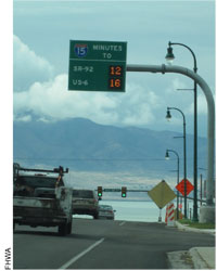 Figure 15. Photo. Hybrid Travel Time Signs for I-15. A photograph shows a travel time electronic sign mounted on a pole arching over a highway lane. It displays travel time in minutes to Route 12 and Route 6. FHWA