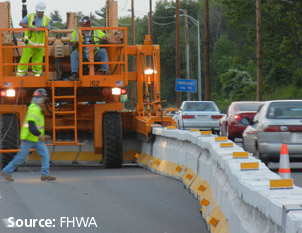 Photo of a zipper barrier and a large mobile machine which adjusts the placement of the zipper barrier. A workers is driving the machine and two other workers are overseeing the work. Cars are positioned on the other side of the zipper barrier.