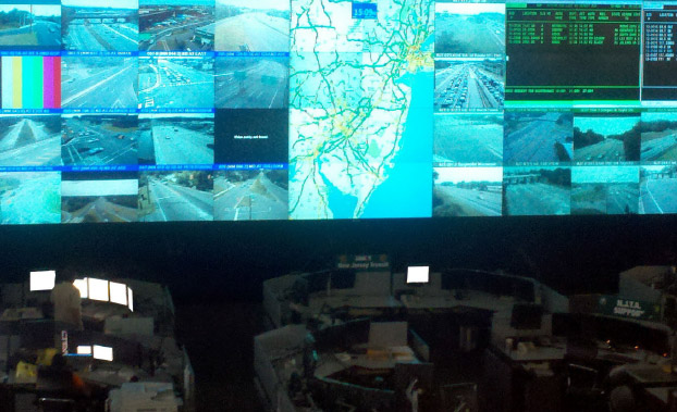 Photo of the inside of the New Jersey Statewide Transportation Management Center. A large screen of monitors in front of several work stations display NJ state mapping, traffic camera feeds, and other data.