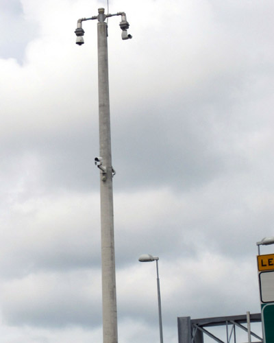 Photo of CCTV cameras for monitoring traffic installed on top of a tall post. Some other highway signs are seen in the corner of the photo. The sky is full of clouds, overcast.