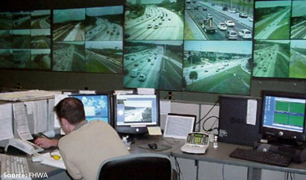 Photo of a dispatcher working inside a TMC siting at a desk/workstation in from of computers, phones and a wall of traffic camera video feeds.