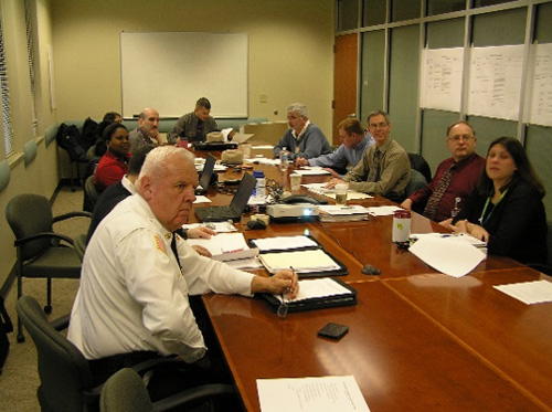 A photo of men and women attending a bi-monthly TIM Steering Committee meeting. Participants are seated at a table in a conference room.