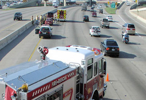 A photo shows a fire engine parked diagonally across the two left lanes of a highway in front of the scene of a traffic incident. Orange cones also divide traffic from the incident.