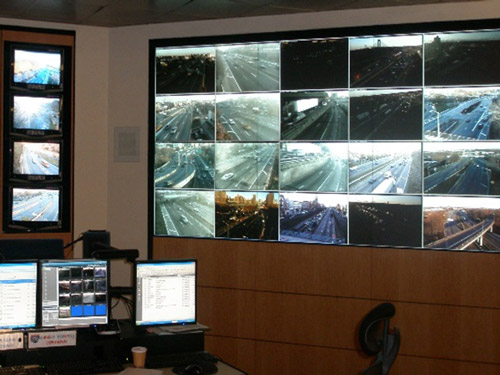 A photo of inside the NYSDOT Joint Traffic Management Center in Queens, NY. Computer screens and several live traffic camera feeds are displayed on a large wall/screen in the room.