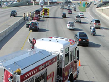 A photo shows a fire engine parked diagonally across the two left lanes of a highway in front of the scene of a traffic incident. Orange cones also divide traffic from the incident.
