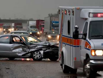 A photo of a car accident scene at the side of the highway. An emergency vehicle sits in front of a heavily damaged passenger car. Highway traffic is seen backed up behind the accident. 
