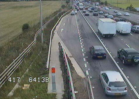 Photo of emergency turnoff on the left side of a freeway in the UK. The turnoff, which is separated from the part-time shoulder lane by a dashed white line, is several hundred feet in length and is wide enough to fit two vehicles side-by-side. A taper is present at the beginning and end of the turnoff.