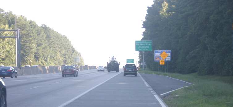 Photo of an emergency turnoff on the right side of a freeway in Georgia. The clear zone just upstream of the turnoff contains a post-mounted sign with the text “Notice” in black text on a yellow background at the top, followed by “Emergency Pull-Off” in black text on a white background and a black arrow pointing upward and to the right directly below. The turnout is separated from the part-time shoulder lane by a dashed white line. A taper is present at the beginning and end of the turnoff.