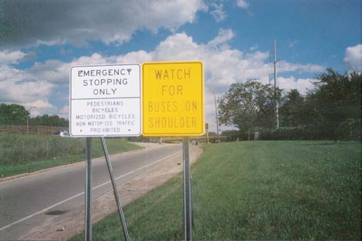 Photo of two adjacent post-mounted signs for BOS operations in Minnesota. The left-sign is a regulatory-series sign stating, “Emergency Stopping Only; Pedestrians, Bicycles, Motorized Bicycles, Non-Motorized Traffic Prohibited” in black text on a white background. The right-sign is a warning-series sign stating, “Watch for Buses on Shoulder” in black text on a yellow background.