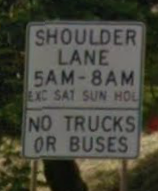 Photo of a post-mounted regulatory sign with black text on a white background. The sign states, “ Shoulder lane 5AM through 8AM Exc Sat Sun Hol, No Trucks or Buses”.