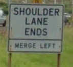 Photo of a post-mounted regulatory sign with black text on a white background. The sign states, “Shoulder lane ends, Merge Left”.