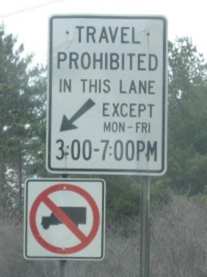 Photo of a post-mounted regulatory sign with black text on a white background. The sign states, “Travel Prohibited in this lane except Mon through Fri 3:00 through 7:00 PM”.
