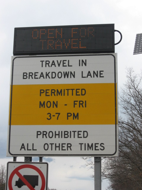 Photo of dynamic message sign added to static regulatory sign below. The dynamic sign mounted on top reads, “Open for travel”, while the static sign below reads, “Travel in breakdown lane, Permitted Mon through Fri 6 through 10 AM 3 through 7 PM, Prohibited all other times”.