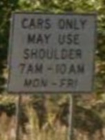 Photo of a post-mounted regulatory sign with black text on a white background. The sign states, “Cars only may use shoulder 7AM through 10AM Mon through Fri”.