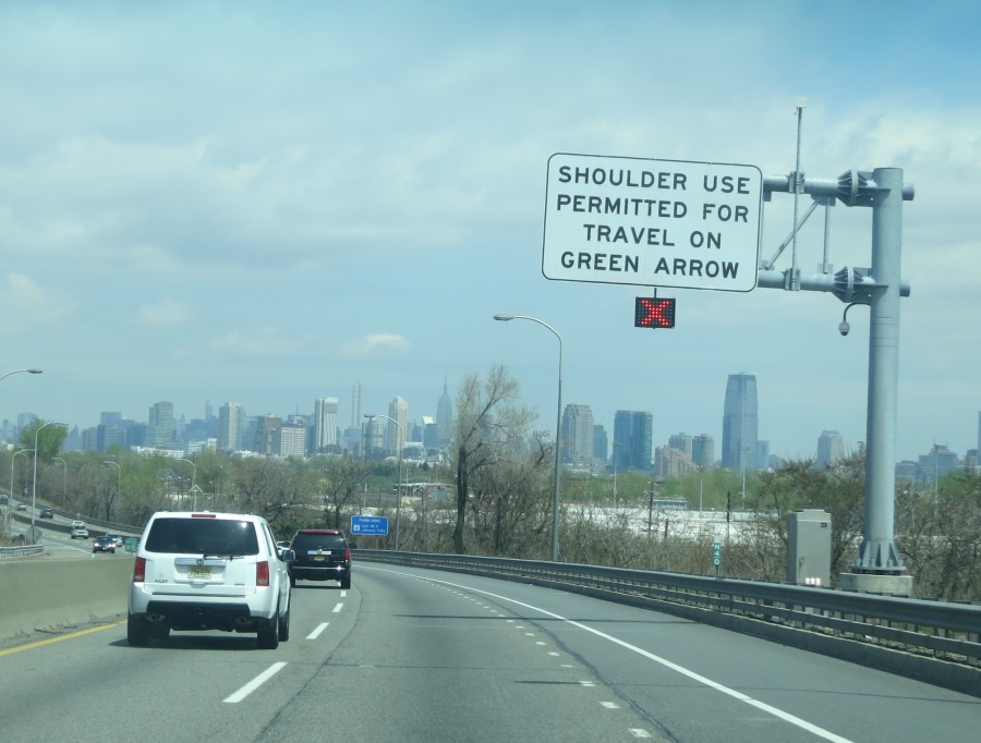 Photo of a partial gantry over the shoulder lane. The regulatory sign with black text on a white background reads, “Shoulder use permitted for travel on green arrow.” Below the static regulatory sign is s dynamic sign displaying a steady red ‘X’.