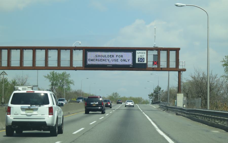 Photo of dynamic sign mounted over the travel lanes on N.J. Turnpike indicating via text the “shoulder [is] for emergency use only” and the variable speed limit, currently set at 50 m.p.h. A dynamic lane-control sign is mounted over the shoulder lane and is displaying a steady red ‘X’ to indicate the shoulder is currently closed.