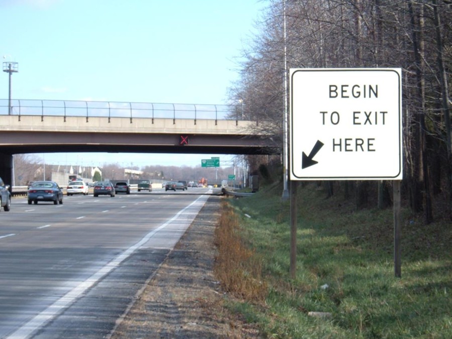 Photo of a static roadside regulatory sign placed at the point where vehicles are allowed to utilize the outside lane (shoulder lane) upstream of an exit ramp. The sign reads “Begin to exit here” in black text on a black background with an arrow point pointing downward and to the left (i.e., towards the shoulder lane).