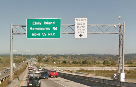 Photo of regulatory sign mounted on an overhead gantry above a shoulder travel lane in Washington State. The sign reads, “Shoulder Open to Traffic, Mon-Fri, 3-7PM” in black text on a white background with an arrow point downward in the direction of the shoulder lane.