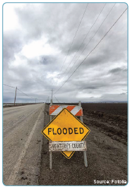 A warning sign mounted on a Type I barricade placed to the right of a rural roaday. The warning sign reads "Flooded". Source: Fotolia