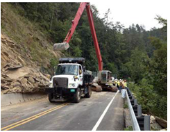 Maintenance crews use bucket loaders and dump trucks to clear a landslide that blocks a roadway.