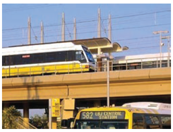 A rail car stopped at a light rail station on a raised platform. Below the platform, a bus is picking up riders.