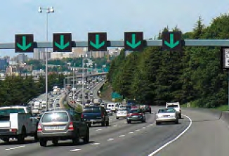 The image shows an overhead gantry with dynamic signs that currently display a green arrow above each lane, indicating that the lanes are open to traffic.